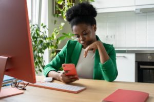A woman procrastinating on her phone while sitting in front of a computer.